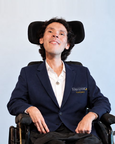 Thiago, responsável pelo pós-venda da Viaggiamo, em sua cadeira de rodas, de blazer azul-marinho com o logotipo da agência.